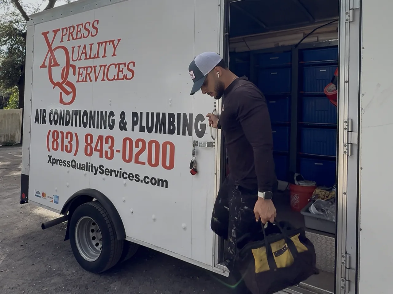 XQS technician carrying a tool bag from an XQS air conditioning and plumbing service trailer